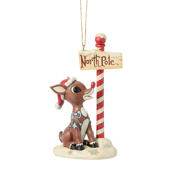 Front view of licensed Rudolph by North Pole Sign Ornament featuring Rudolph seated beside a North Pole sign on a snowy base.