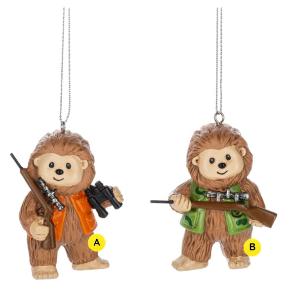Two Bigfoot Hunting ornaments shown together, each featuring a smiling Bigfoot holding a hunting rifle, with one wearing an orange safety vest and carrying binoculars and the other wearing a green camo-style vest.