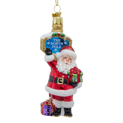 Glass Santa at the North Pole ornament with Santa holding a green gift beside a candy cane striped North Pole sign and purple present.