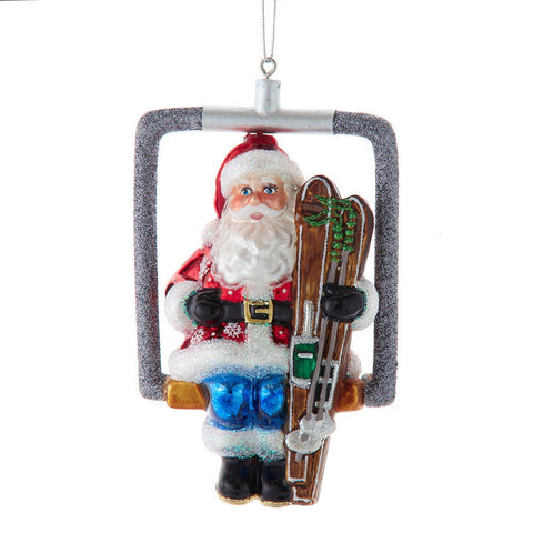 Glass Santa ski lift ornament featuring Santa in a silver glitter chair lift, holding brown skis, and wearing a red coat, blue pants, and black boots.