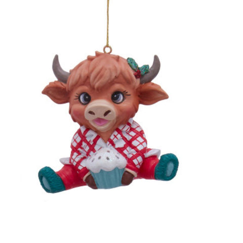 Highland cow ornament in red plaid pajamas, sitting with a cupcake and wearing a holly accent near one horn.