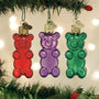Three gummy bear-shaped Christmas ornaments in red, purple, and green hanging on a tree.