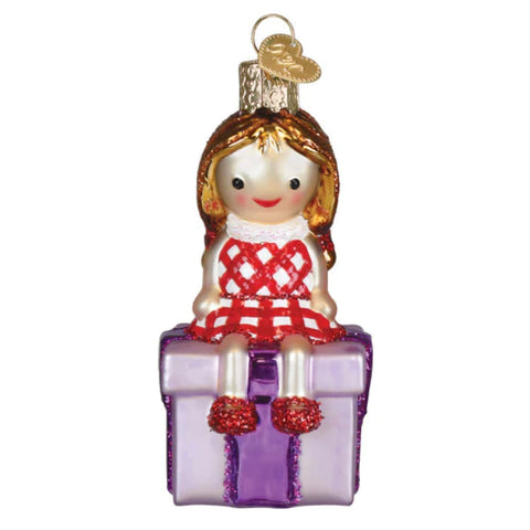 Glass ornament of a girl in a red dress sitting on a purple present against a white background