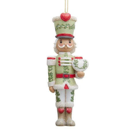 Nutcracker ornament in soft green, red, and cream holding a white heart, with mistletoe detailing and heart accents on the hat and boots.
