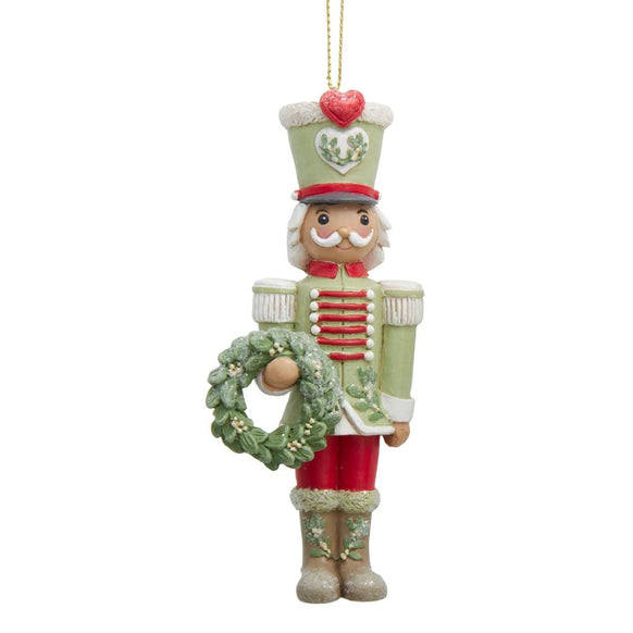 Nutcracker ornament in soft green, red, and cream holding a wreath, with mistletoe detailing and heart accents on the hat and boots.