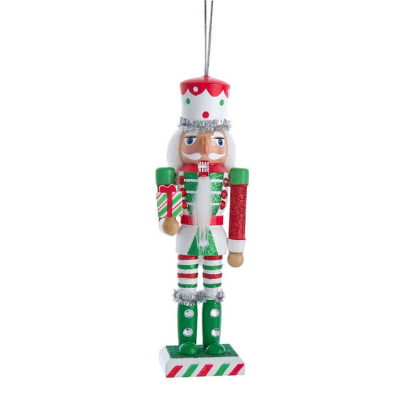 Red, green, and white nutcracker ornament holding a striped present, with glittered details, white beard, and silver tinsel trim on a rectangular base.