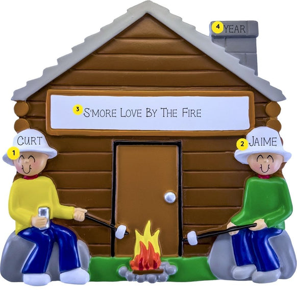 Personalized camping cabin couple ornament featuring two people roasting marshmallows by a campfire outside a brown cabin. The design includes two names, a custom phrase on the cabin, and a year on the chimney.