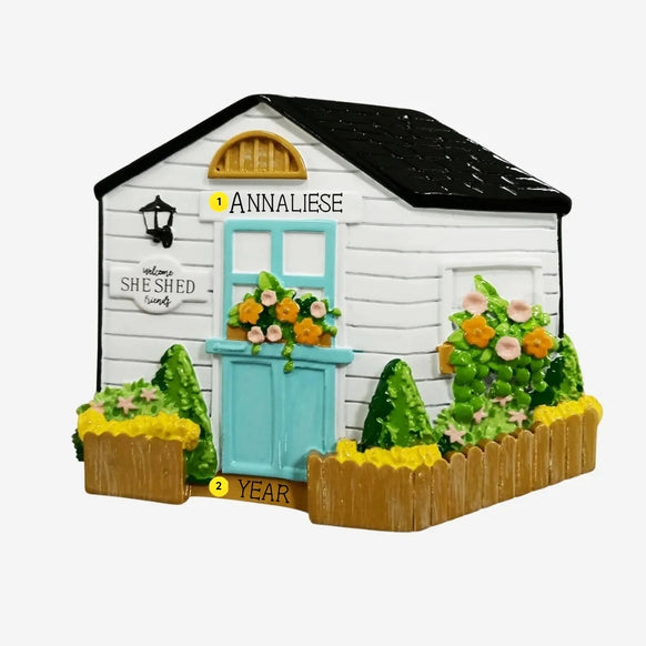 Personalized she shed garden cottage ornament with a white exterior, black roof, aqua door, flower boxes, and name and year personalization.