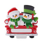 Snowman couple ornament riding in a red holiday truck with festive hats, scarves, and mittens.