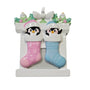 Twin penguin stocking ornament with one pink stocking and one blue stocking hanging from a white fireplace mantel with holly, candles, and a bow.