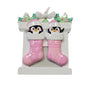 Twin penguin stocking ornament with two pink stockings hanging from a white fireplace mantel decorated with holly, candles, and a bow.
