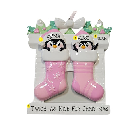 Personalized twin penguin stocking ornament with two pink stockings, two baby names on the hats, a year on the mantel, and the phrase “Twice As Nice For Christmas” on the base.
