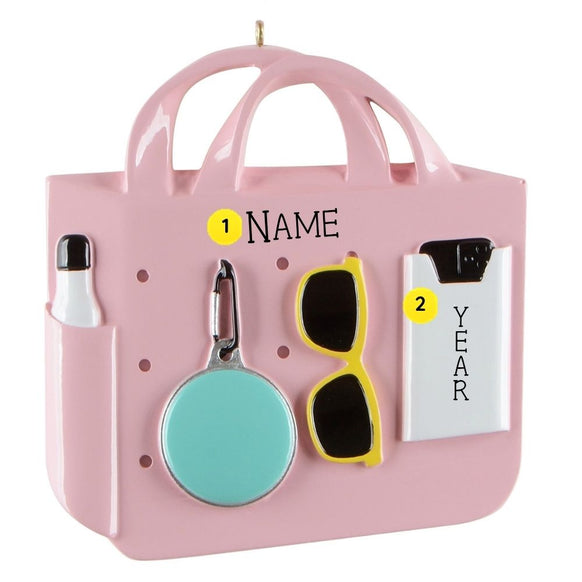 Personalized pink beach bag ornament with a white bottle, aqua round accessory, yellow sunglasses, and white clipboard showing name and year placement.