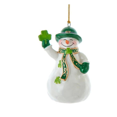 White snowman ornament wearing a green hat and scarf, holding a shamrock, with green accents and a gold hanging cord.