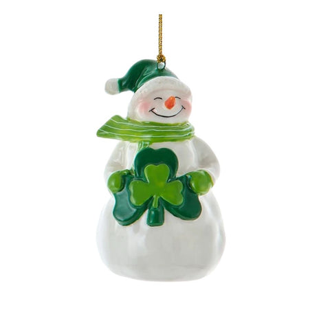 White snowman ornament wearing a green cap and striped scarf, holding a green shamrock at its chest with a gold hanging cord.