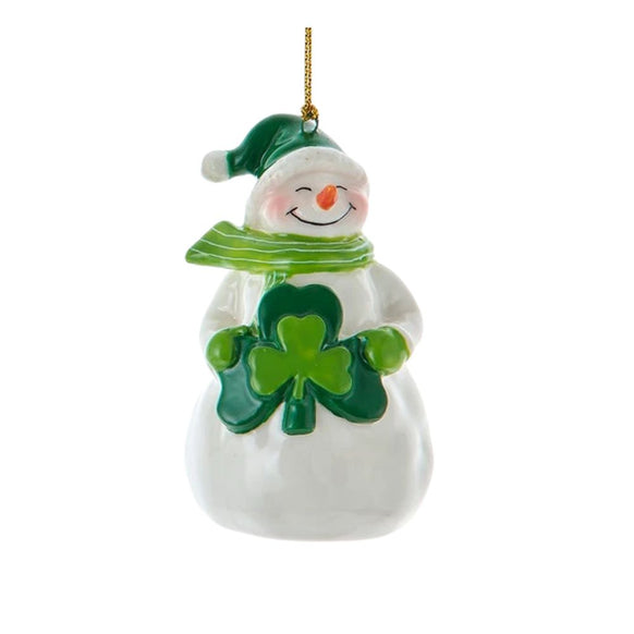 White snowman ornament wearing a green cap and striped scarf, holding a green shamrock at its chest with a gold hanging cord.
