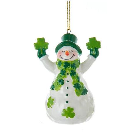 White snowman ornament wearing a green top hat and shamrock scarf, holding shamrocks in both hands with shamrock accents on its body.
