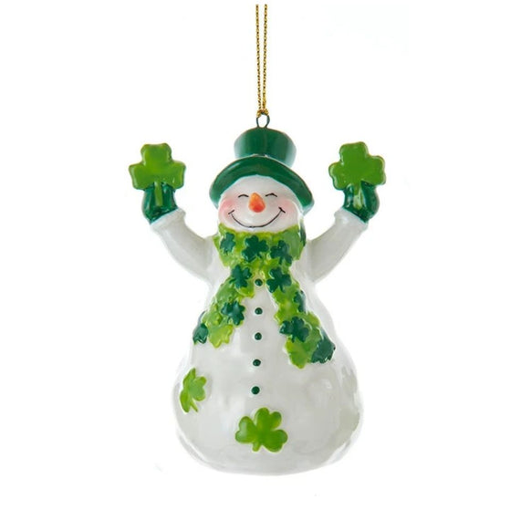 White snowman ornament wearing a green top hat and shamrock scarf, holding shamrocks in both hands with shamrock accents on its body.