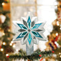 Silver, white, and blue snowflake tree topper displayed on top of a decorated Christmas tree branch with lights in the background.