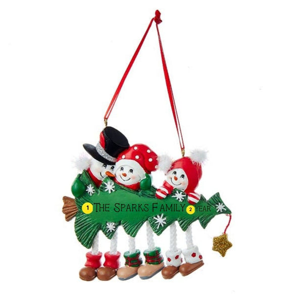 Personalized snowman family of 3 ornament with three smiling snowmen peeking over a decorated Christmas tree with dangling legs and space for a family name or short phrase and year.