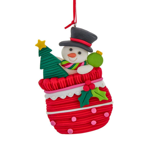 Snowman ornament peeking out of a red gift bag with holly detail, holding a green ornament beside a Christmas tree.