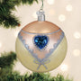 Decorative Christmas ball ornament with blue heart and silver glitter on a blurred Christmas tree background.