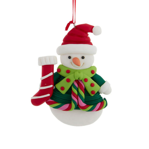 Snowman ornament wearing a red hat and green scarf with red dots, holding a red Christmas stocking with candy cane trim.