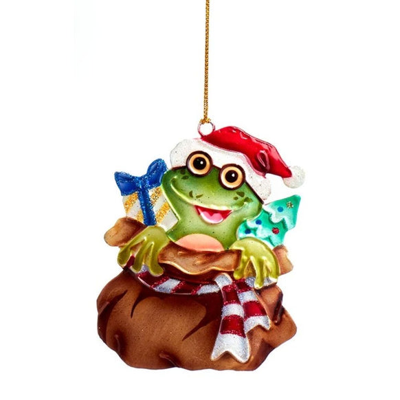 Tin frog ornament wearing glasses and a Santa hat, sitting in Santa’s gift sack with a striped scarf, present, and tiny tree.