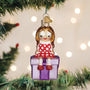 Christmas ornament of a girl sitting on a present under a decorated tree with lights.