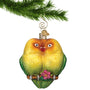 Glass Lovebirds Ornament sitting together in yellows, orange and green with pink flower hanging from a gold swirl hook from a Christmas tree branch